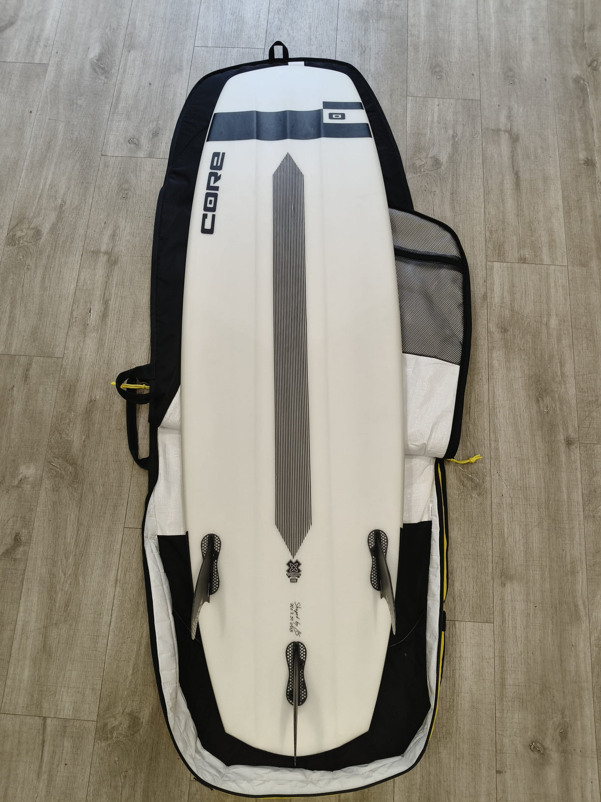 A white surfboard with black and gray graphics, resting inside a black and gray soft-sided carrying bag on a light wood floor.