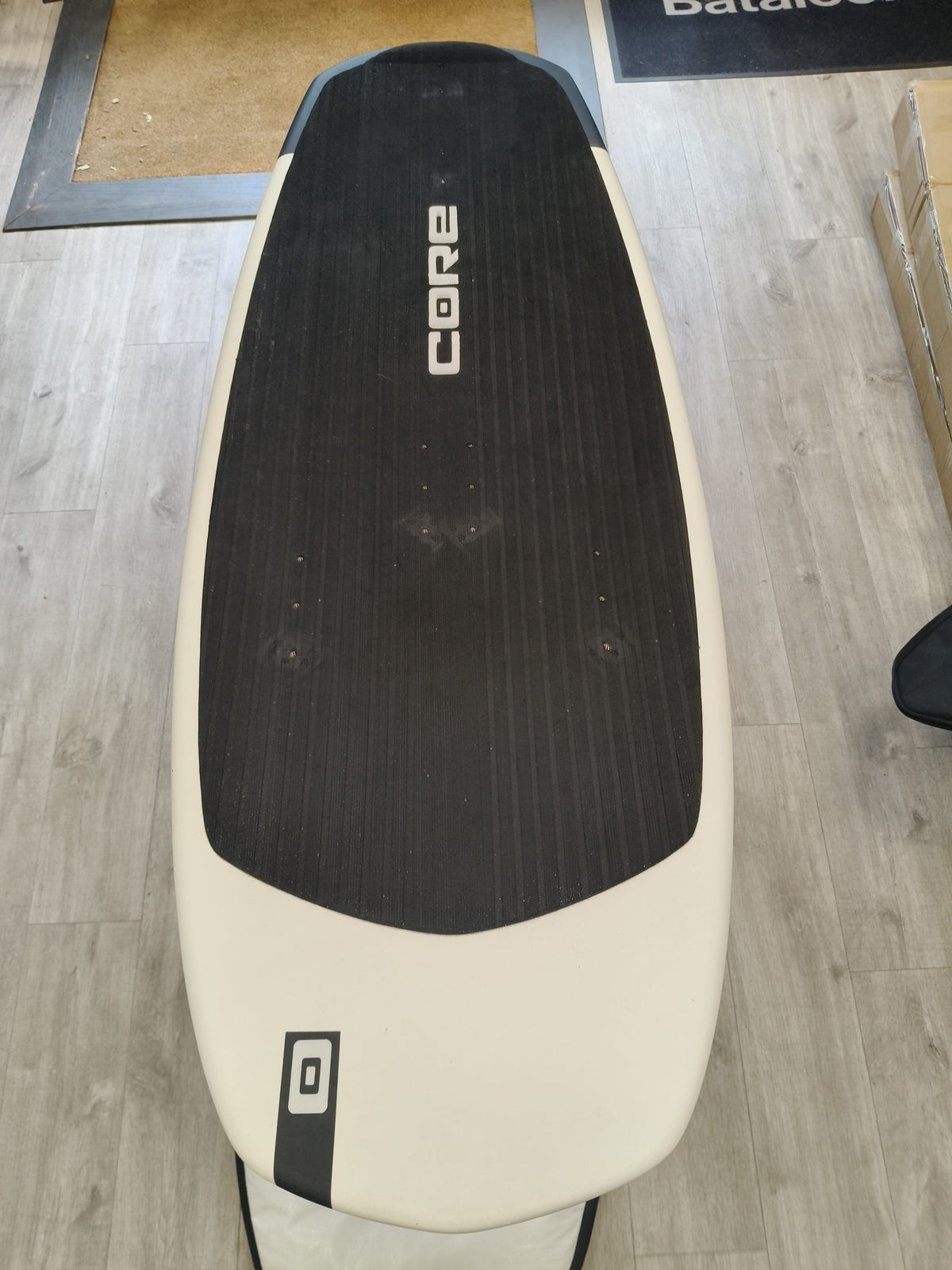 A CORE brand stand-up paddleboard shown from a top-down perspective on a light wood floor. The board features a cream-colored lower section with a black graphic accent and a black textured grip pad on top.