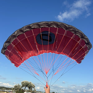 A red and black parachute canopy with red and blue suspension lines, being held up by a person's hand against a blue sky with clouds.