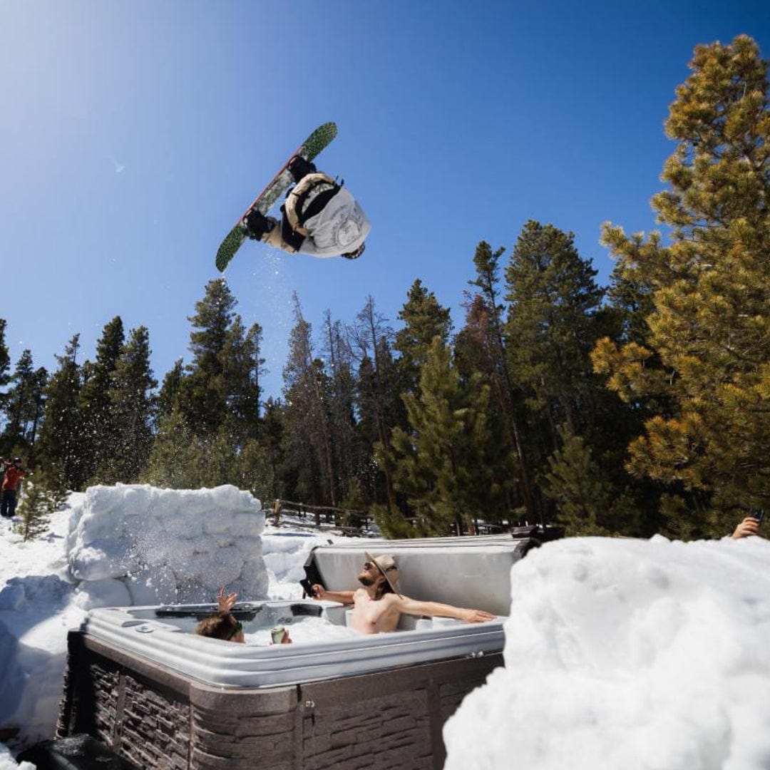 A person snowboarding and jumping over a hot tub in a snowy landscape with trees.