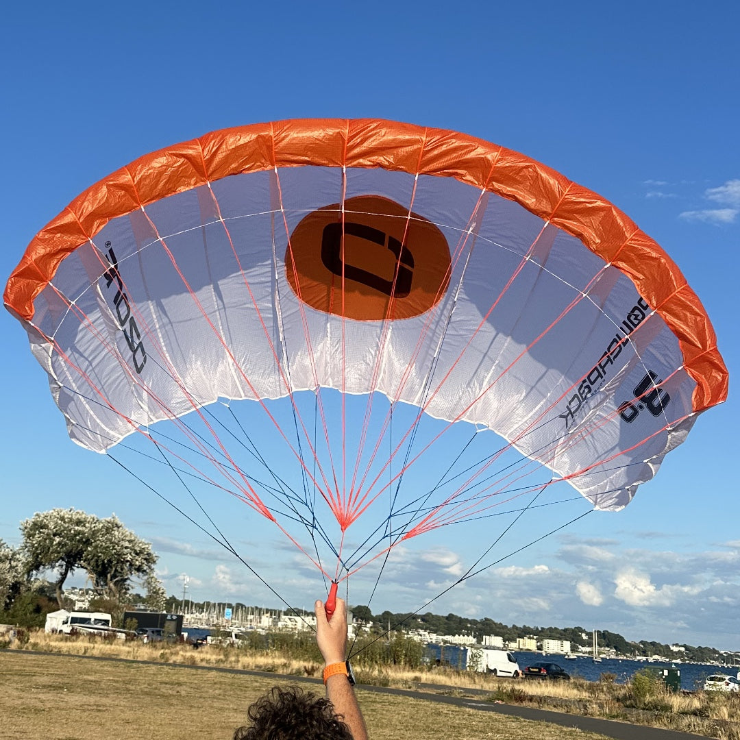 An orange and white parachute is being held up by a person outdoors under a blue sky, with a beach and boats visible in the background.