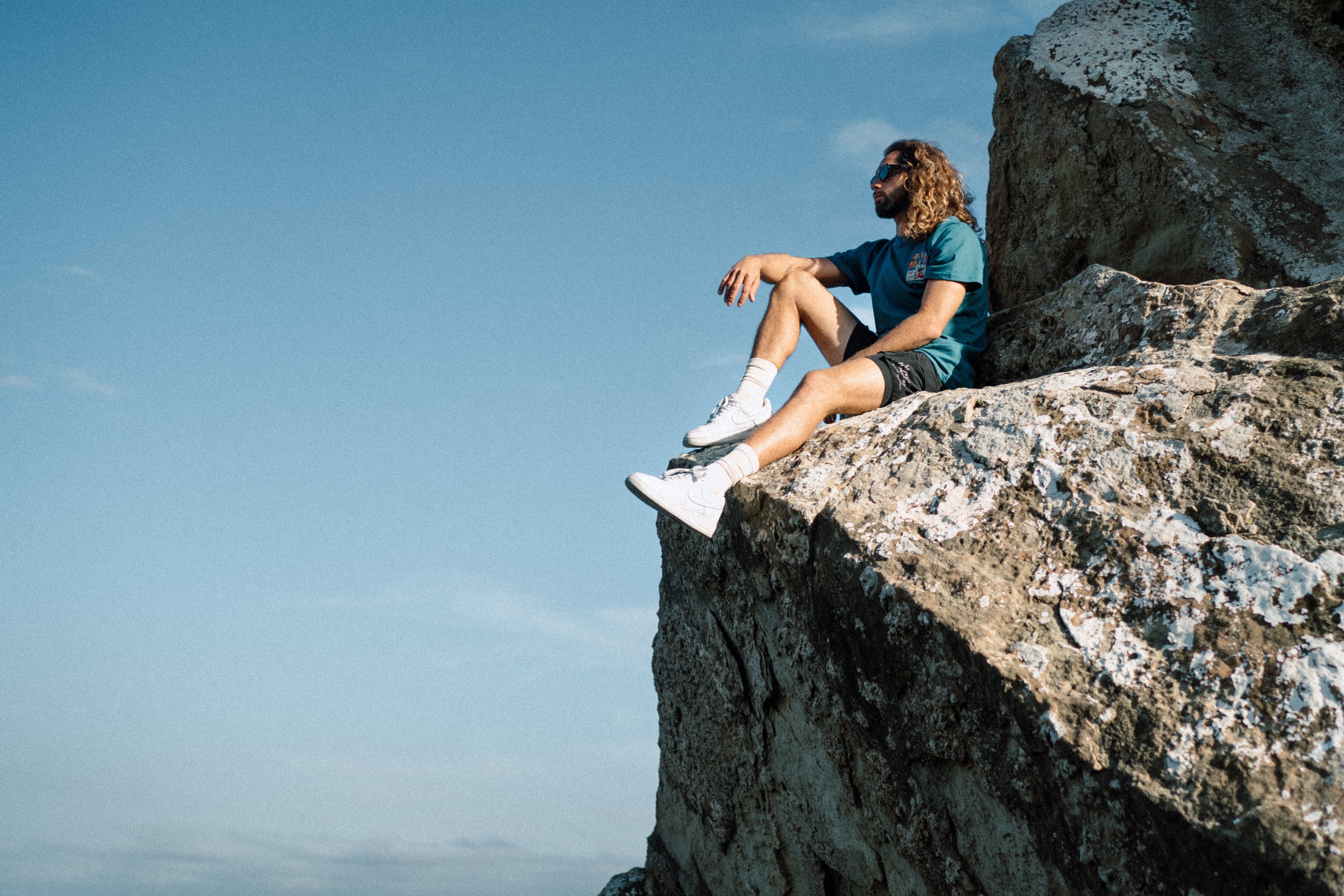 Person sitting on a rocky outcrop with a clear blue sky, wearing S2AS clothing